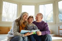 Grandparents on window seat reading book with grandson, smiling — Stock Photo