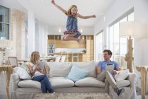 Menina pulando no ar do sofá da sala de estar, enquanto os pais usando tablet digital — Fotografia de Stock