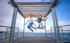 Due giovani amiche che saltano all'unisono sul molo del mare — Foto stock