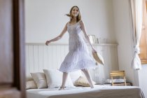 Молода жінка в довгій білій сукні стрибає на ліжку дивиться на камеру посміхаючись — стокове фото