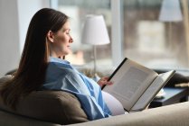 Pregnant woman sitting on sofa, reading book — Stock Photo