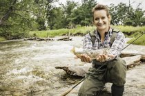 Woman crouching in river holding freshly caught fish looking at camera smiling — Stock Photo