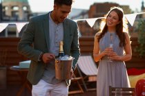Casal adulto médio na festa no terraço do telhado segurando balde de gelo com champanhe sorrindo — Fotografia de Stock