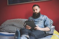 Giovane uomo barbuto utilizzando tablet digitale sul letto — Foto stock