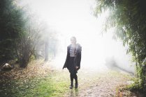 Повнометражний вид спереду молода жінка стоїть в туманному саду, дивлячись на камеру посміхаючись — стокове фото