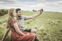 Молодая пара, сидящая на вершине холма, делает селфи со смартфоном, Коди, Вайоминг, США — стоковое фото