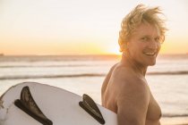 Чоловік на пляжі носить дошку для серфінгу, дивлячись через плече на камеру посміхаючись — стокове фото