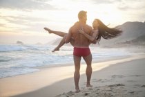Середня доросла пара на пляжі, чоловік носить жінку в руках, вид ззаду — стокове фото