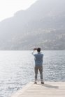 Rear view of young man taking photograph from pier, Lake Mergozzo, Verbania, Piemonte, Italy — Stock Photo