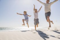 Father and sons on beach, arms raised jumping in mid air — Stock Photo