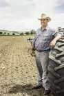 Portrait of male farmer leaning against tractor tyre in ploughed field — Stock Photo