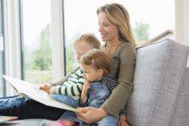 Mother and two young children sitting on sofa reading a book — Stock Photo