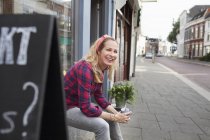 Donna seduta sul davanzale del negozio con in mano una tazza di caffè che guarda altrove sorridendo — Foto stock