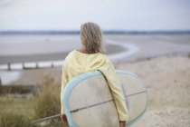 Mujer mayor caminando hacia la playa, llevando tabla de surf, vista trasera - foto de stock