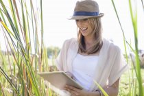 Mitte erwachsene Frau sitzt im langen Gras und benutzt ein digitales Tablet — Stockfoto