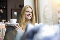 Молода жінка з довгим світлим волоссям сидить тримаючи чашку кави, дивлячись в очі посміхаючись — стокове фото