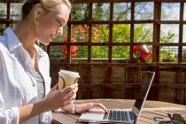Молодая женщина с кофе на вынос и ноутбук в саду — стоковое фото