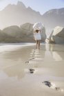 Pegadas e visão traseira da mulher na praia vestindo vestido branco curto segurando guarda-chuva — Fotografia de Stock