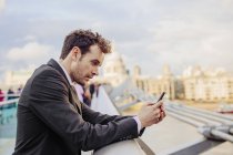 Empresário lendo mensagem de texto do smartphone enquanto se inclina na ponte Millennium, Londres, Reino Unido — Fotografia de Stock