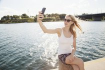 Jovem mulher sentada à beira do rio levando selfie smartphone, Ilha do Danúbio, Viena, Áustria — Fotografia de Stock