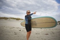 Старшая женщина стоит на пляже, держа доску для серфинга, указывая на море — стоковое фото