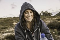 Mujer con capucha superior sosteniendo botella de agua mirando hacia abajo sonriendo - foto de stock