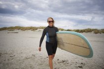 Mujer mayor caminando en la playa, llevando tabla de surf - foto de stock