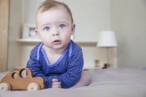 Portrait of blue eyed baby boy sitting up on bed with wooden toy car — Stock Photo