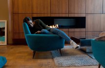 Woman in armchair in front of wooden cabinets and open fire using smartphone — Stock Photo