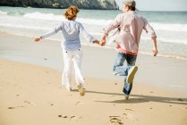 Зрелая пара, держащаяся за руки, бегущая по пляжу, Камаре-сюр-мер, Бретань, Франция — стоковое фото