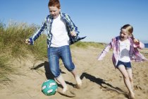 Menina e menino perseguindo futebol através da areia, sorrindo — Fotografia de Stock