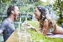 Young man in river, young woman lying on grass at edge of river, man splashing woman with water — Stock Photo