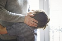 Father and son hugging by window indoors, boy looking up — Stock Photo