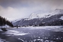 Zugefrorener See und Dorf unter schneebedecktem Berg, Engadin, Schweiz — Stockfoto