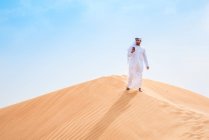 Hombre de Oriente Medio que usa ropa tradicional usando teléfono inteligente en la duna del desierto, Dubai, Emiratos Árabes Unidos - foto de stock