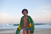 Jeune homme sur la plage, enveloppé dans le drapeau brésilien — Photo de stock