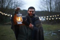 Young male camper holding up lantern whilst wrapped in blanket in woods — Stock Photo