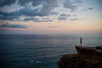 Женщина, стоящая на скале и глядящая в море на закате — стоковое фото