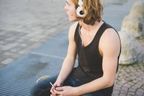 Junger Mann sitzt auf Gehweg und hört Kopfhörermusik — Stockfoto