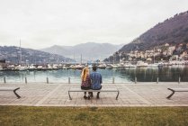 Vista trasera de la joven pareja sentada en el banco con vistas al lago de Como, Italia - foto de stock