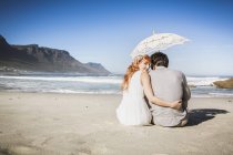 Rückansicht einer Frau mit Arm um den Mann am Strand sitzend, Regenschirm haltend, über die Schulter in die Kamera blickend — Stockfoto