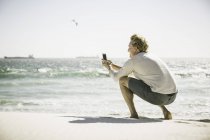 Homme mûr accroupi sur la plage prenant des photos de la mer, à l'aide d'un smartphone — Photo de stock