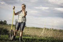 Mujer en la granja sosteniendo pala usando un teléfono inteligente para hacer una llamada telefónica - foto de stock