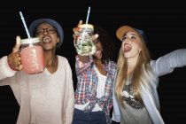 Mujeres jóvenes sosteniendo frascos de albañil brazos levantados con la boca abierta sonriendo - foto de stock