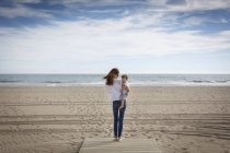 Vista trasera de la mujer que lleva a su hija pequeña en la playa, Castelldefels, Cataluña, España - foto de stock