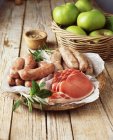 Plate of raw bacon and sausages with wicker basket of apples — Stock Photo