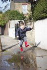 Criança masculina em botas de borracha vermelha pulando na poça da calçada — Fotografia de Stock