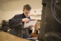Impresora masculina joven inspeccionando papel para maquinaria de impresión en taller de impresión - foto de stock