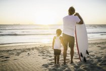 Vater und Sohn stehen am Strand, mit Surfbrett, Blick aufs Meer, Rückansicht — Stockfoto