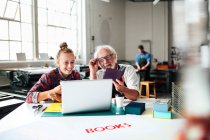 Senior male craftsman laughing and looking at laptop with young woman in book arts workshop — Stock Photo